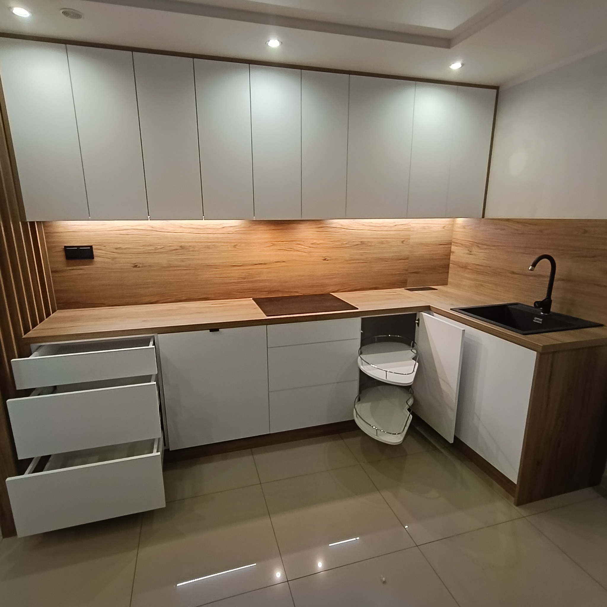 Nowoczesna kuchnia z białymi szafkami i drewnianym blatem. Widoczny wysuwany kosz narożny i otwarte szuflady. Minimalistyczny design z dbałością o detale.