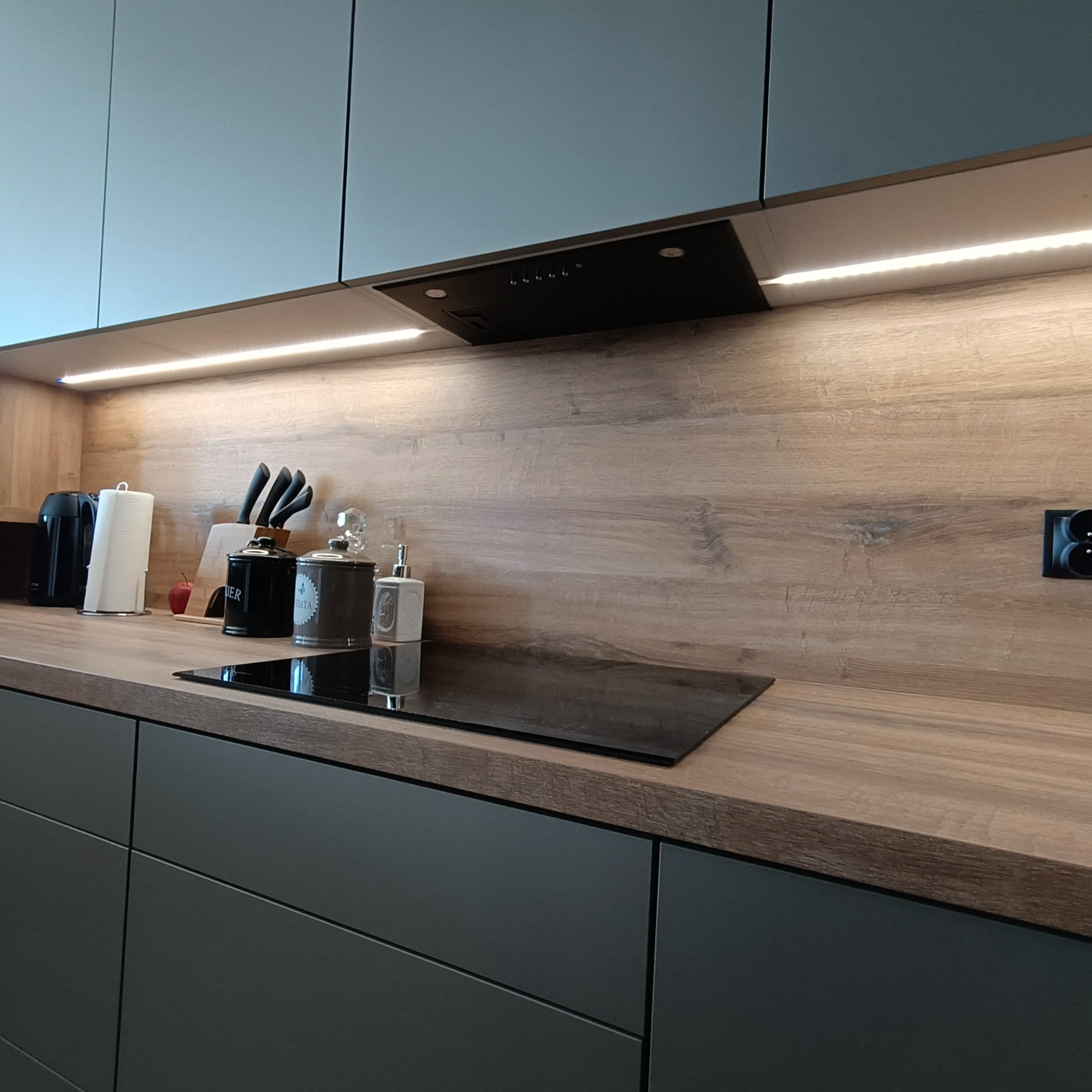 Nowoczesna kuchnia z szarymi szafkami, drewnianym blatem i podświetleniem LED. Na blacie płyta indukcyjna, pojemniki i akcesoria kuchenne.