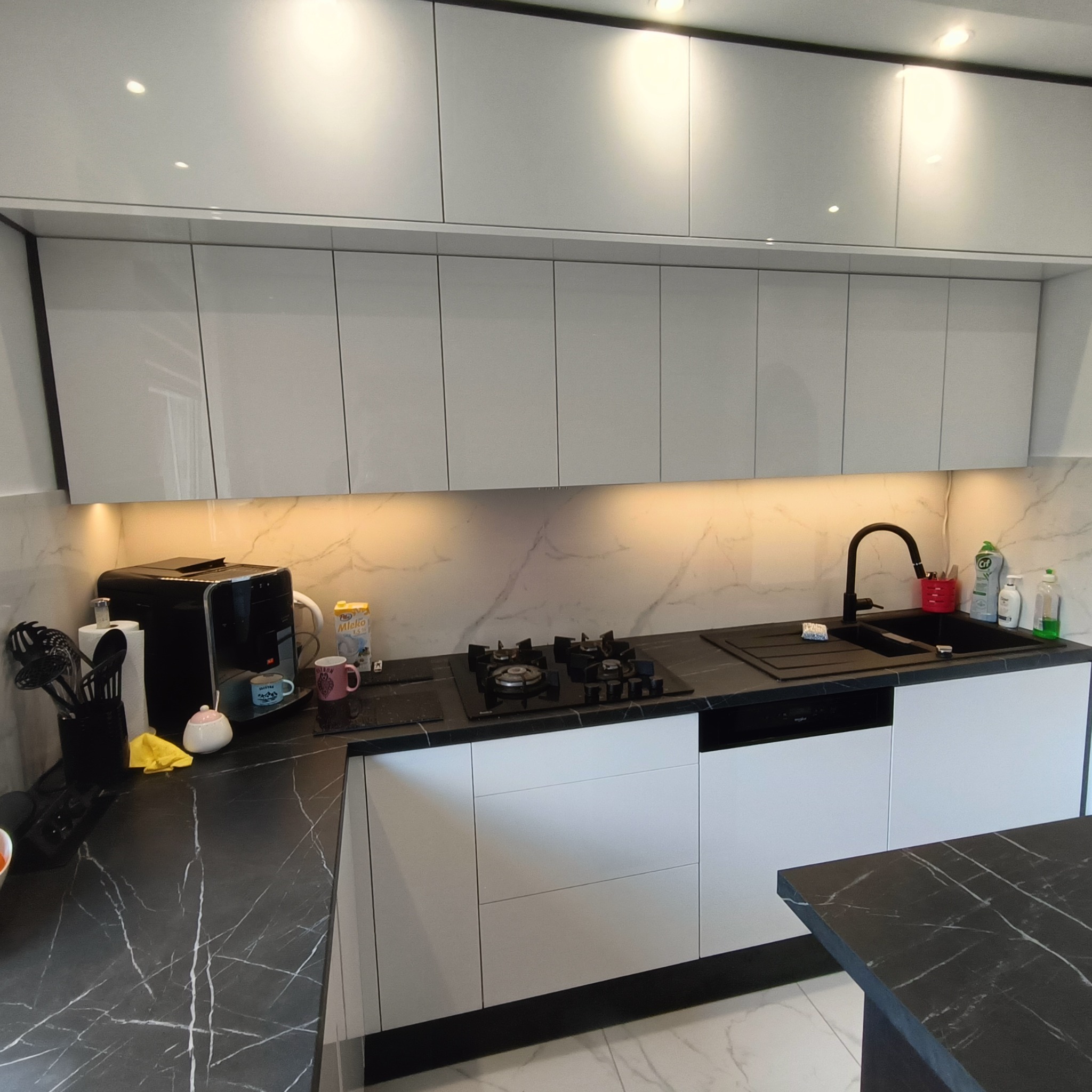 Nowoczesna, jasna kuchnia z czarnym blatem i białymi szafkami, oświetlona punktowo, z marmurowym motywem na ścianie. Widoczny ekspres do kawy i płyta gazowa.