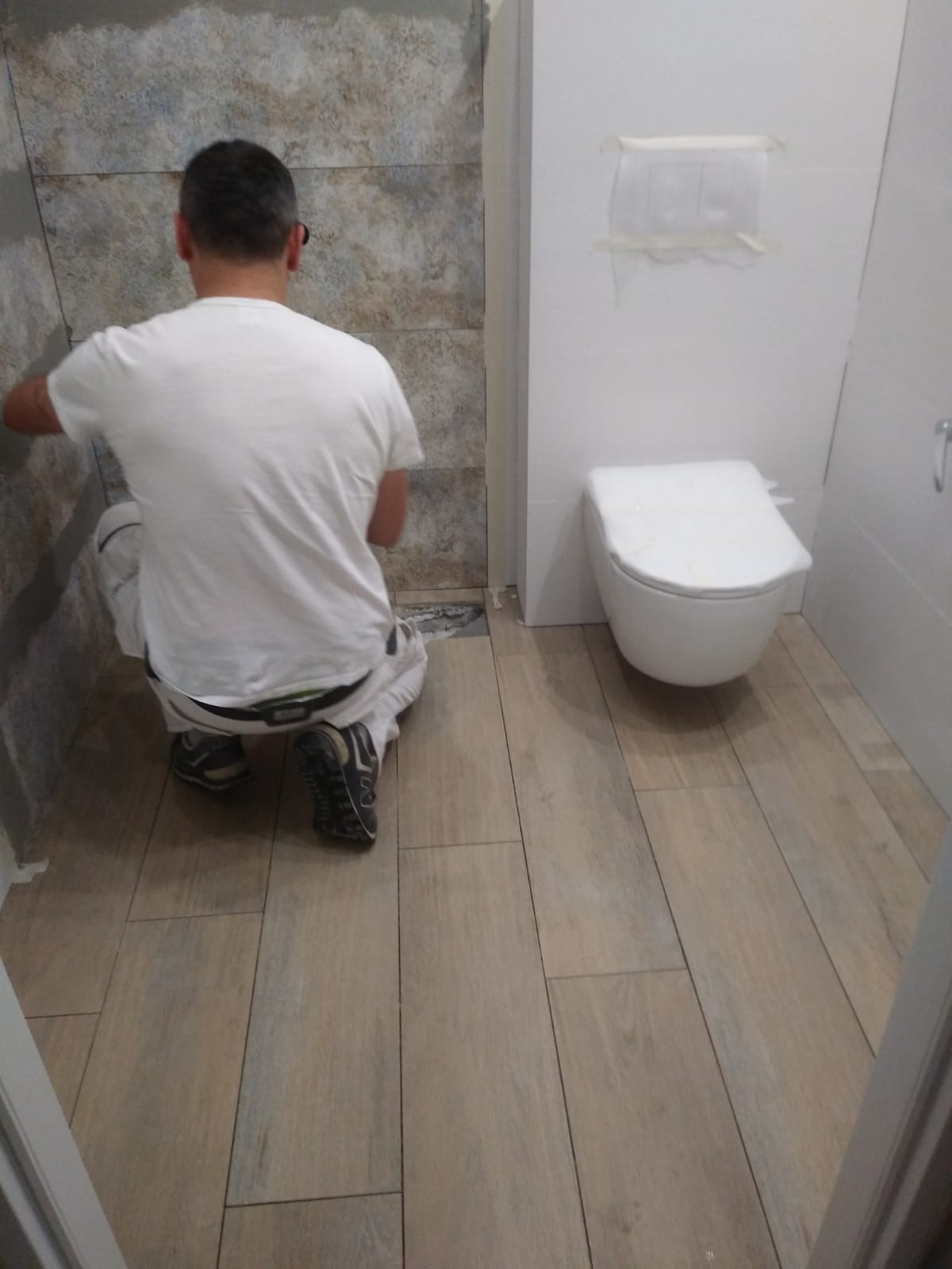 Pracownik w trakcie układania płytek imitujących kamień na ścianie w łazience, widoczna toaleta zabezpieczona folią.