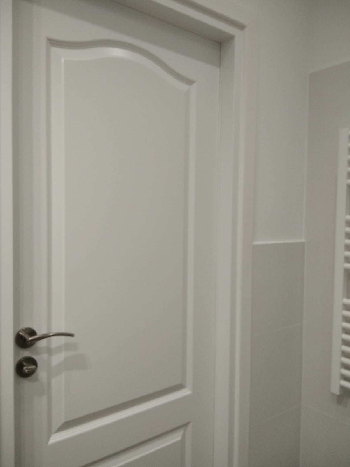 Zamknięte, białe, wewnętrzne drzwi z ozdobną ramą i klamką na tle jasnej ściany z fragmentem grzejnika drabinkowego.