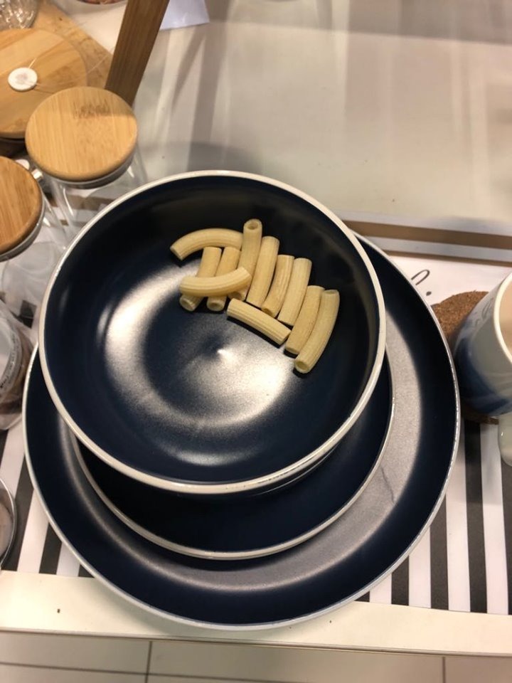 Stos naczyń: granatowa miska z makaronem rurki na granatowym talerzu, ustawione na czarno-białej macie.