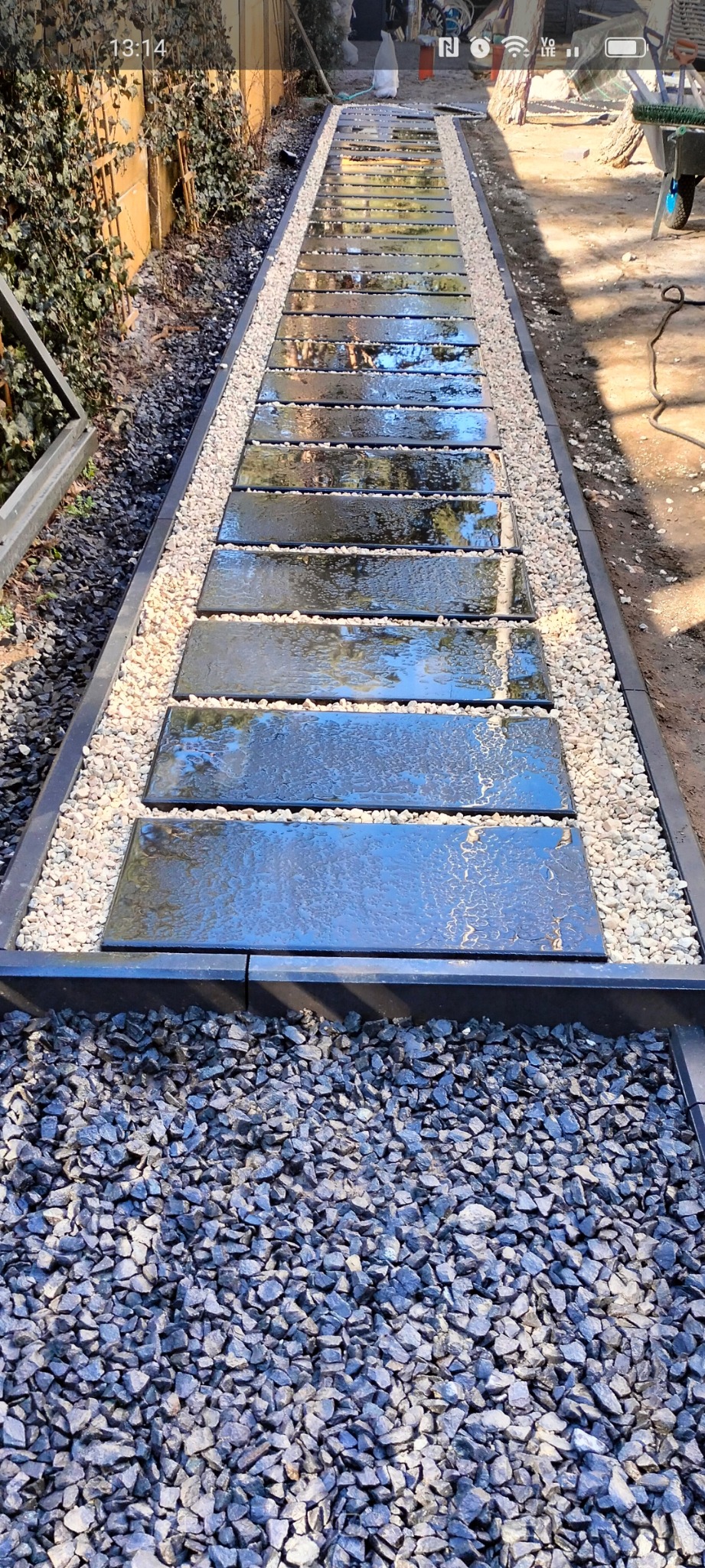 Ścieżka w ogrodzie wykonana z ciemnych, prostokątnych płyt ułożonych w odstępach, otoczona białym i szarym żwirem, z widocznymi refleksami na mokrej powierzchni płyt.