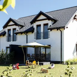 Nowoczesny dom jednorodzinny z białymi ścianami i ciemną dachówką, widoczny z perspektywy ogrodu z zabawkami dziecięcymi na trawniku i meblami ogrodowymi pod parasolem.