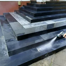 Czyszczenie wodą pod ciśnieniem schodów wyłożonych czarną kostką granitową, widoczna dysza pistoletu natryskowego i strumień wody.