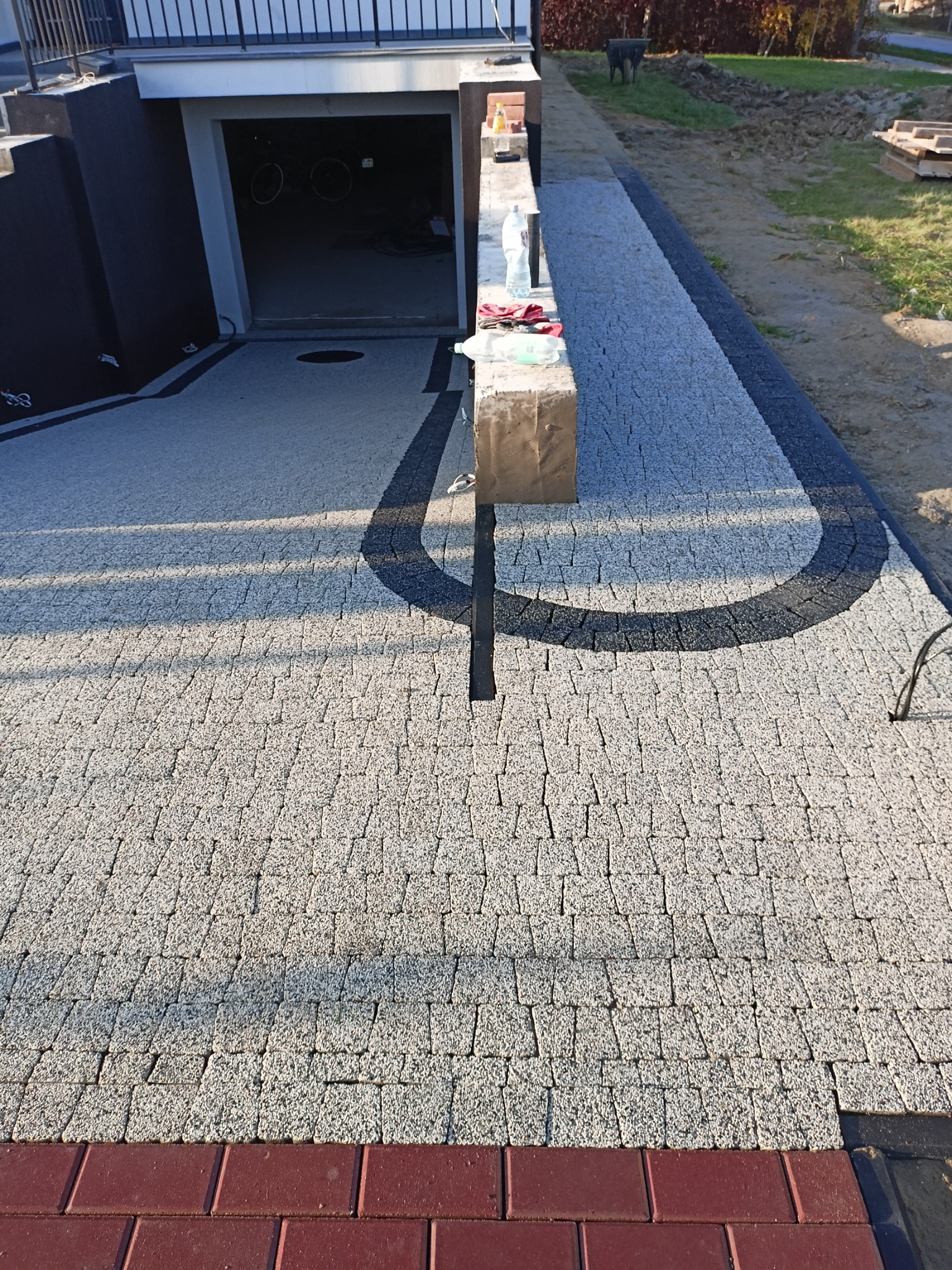 Nowo ułożona nawierzchnia z szarej kostki brukowej z czarnymi akcentami, widoczny wjazd do garażu i przygotowane materiały budowlane