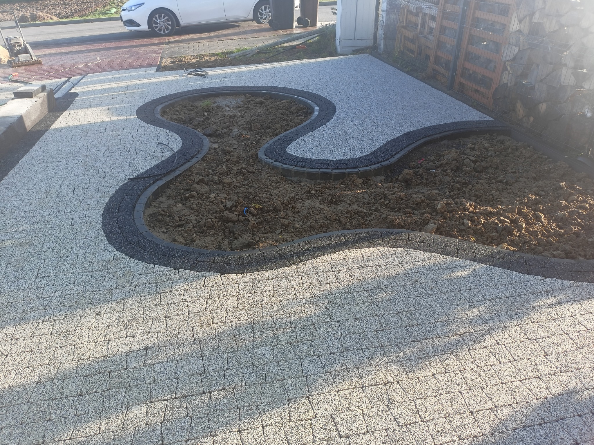 Ułożony chodnik z jasnoszarej kostki brukowej z ciemnoszarym obramowaniem w kształcie falistych rabat, widoczna ziemia wewnątrz rabat, zaparkowany samochód w tle.