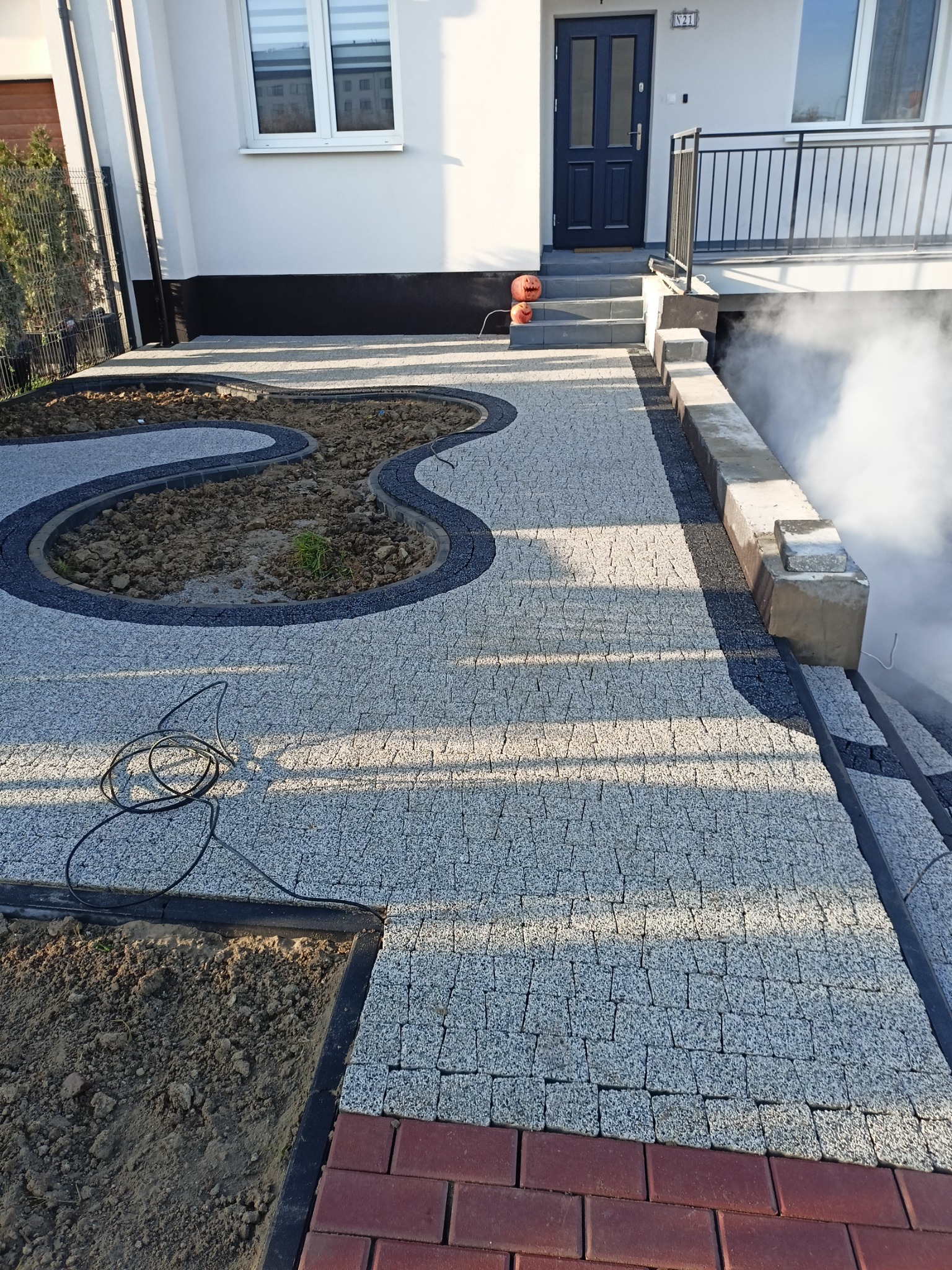 Ułożona szara kostka granitowa przed domem z wyznaczonymi miejscami na rabaty w kształcie falistych linii, obrzeża z ciemnej kostki, widoczne kable i elementy dekoracyjne przy wejściu.