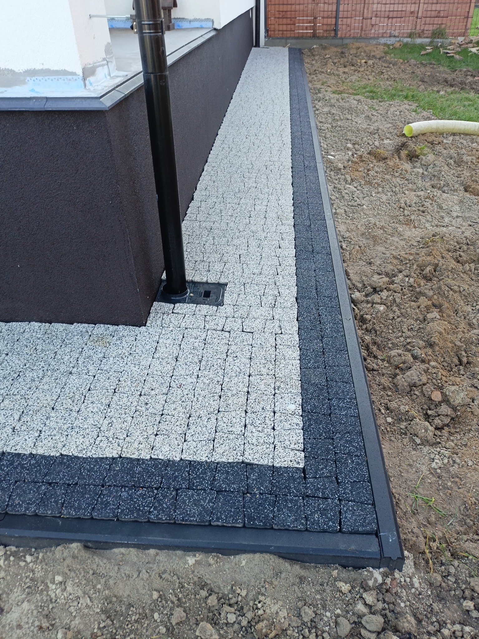 Ułożona ścieżka z jasnoszarej kostki brukowej z czarnym obramowaniem, przylegająca do ściany budynku z rurą spustową. Widoczny fragment terenu przed budynkiem.