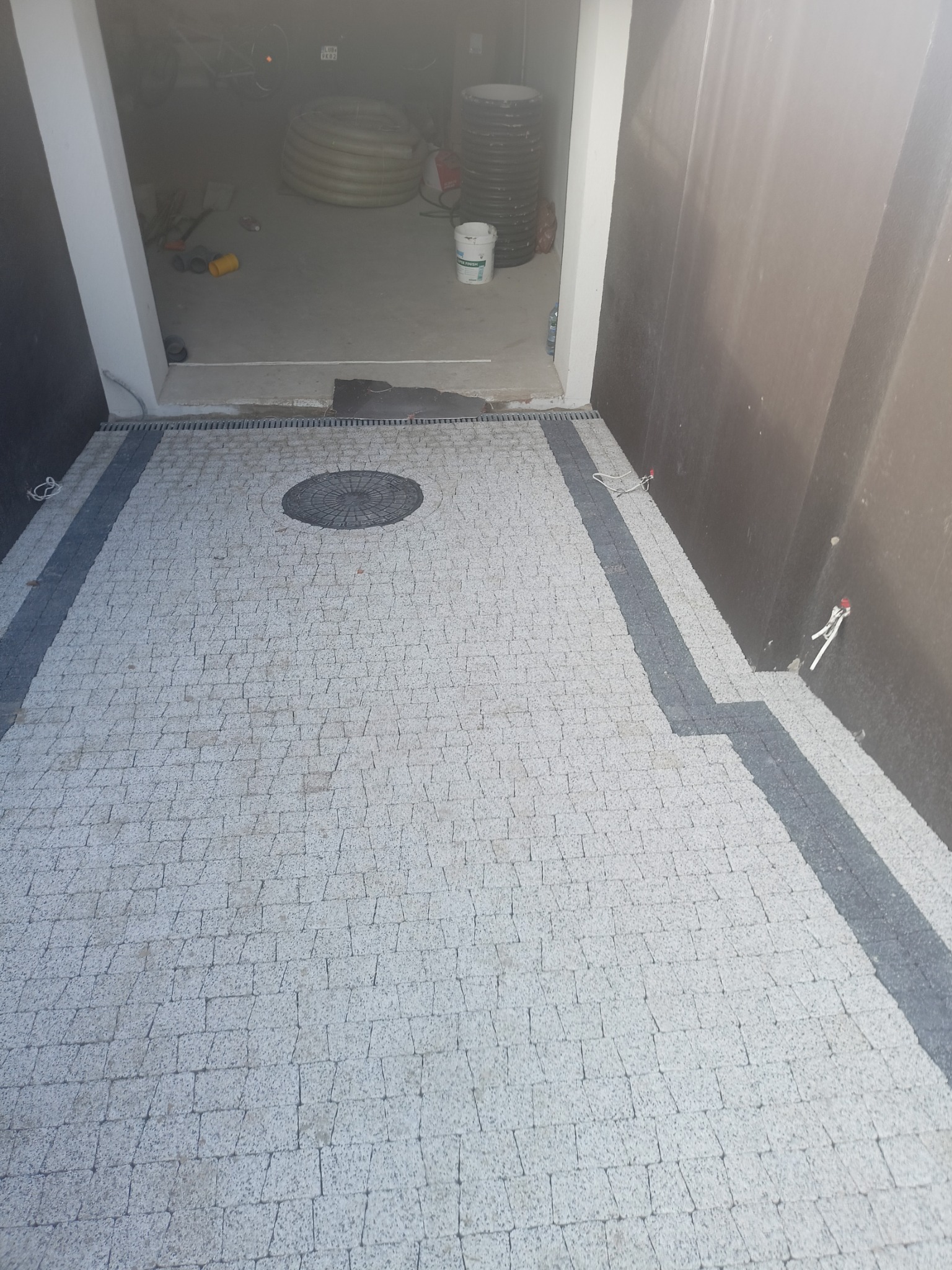 Ułożona jasnoszara kostka brukowa z ciemnoszarymi pasami po bokach i okrągłą studzienką kanalizacyjną na środku, widok z góry, w tle otwarte drzwi garażowe.