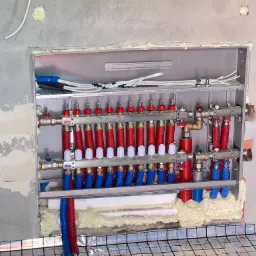 Rozdzielacz ogrzewania podłogowego w trakcie montażu, widoczne rury w kolorach czerwonym i niebieskim, izolacja pianką montażową, folia aluminiowa z siatką oraz surowa ściana w tle.