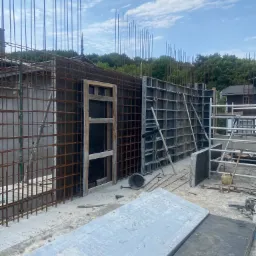 Konstrukcja zbrojeniowa z pionowymi prętami stalowymi wystającymi ponad szalunki, widoczne elementy drewniane i metalowe, niebo z chmurami w tle.