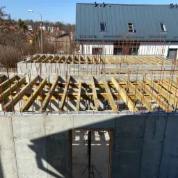 Wysokie fundamenty domu w trakcie budowy, widoczne zbrojenia i drewniane szalunki stropu, w tle budynek z szarą blachą na dachu.