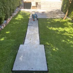 Betonowa ścieżka w ogrodzie z świeżo ułożoną trawą, prowadząca do schodów i wejścia do budynku, z widocznym bębnem z wężem ogrodowym.