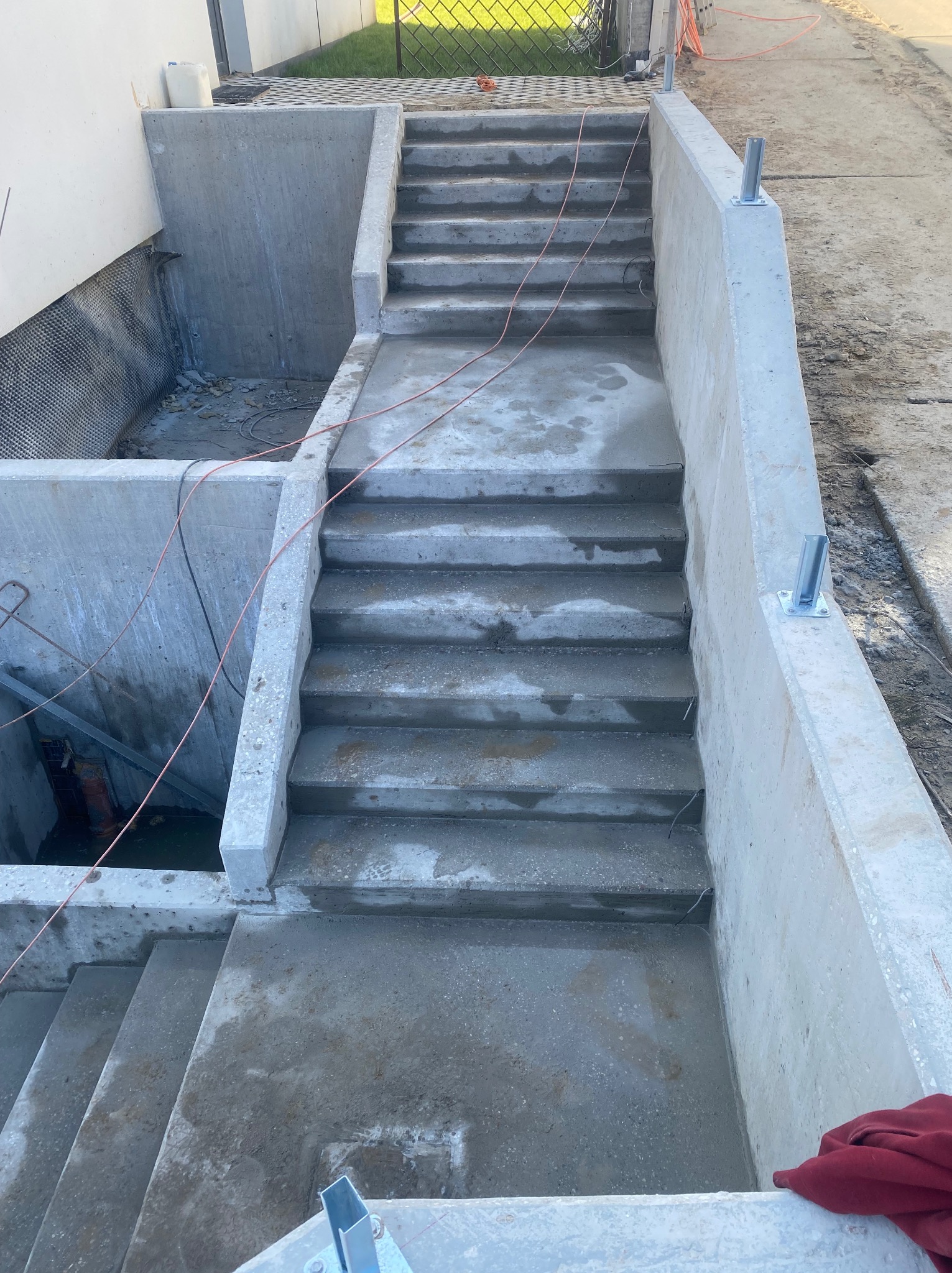 Surowe betonowe schody zewnętrzne z metalowymi uchwytami na balustrady, prowadzące w dół, obok betonowej konstrukcji z hydroizolacją. Widok z góry.