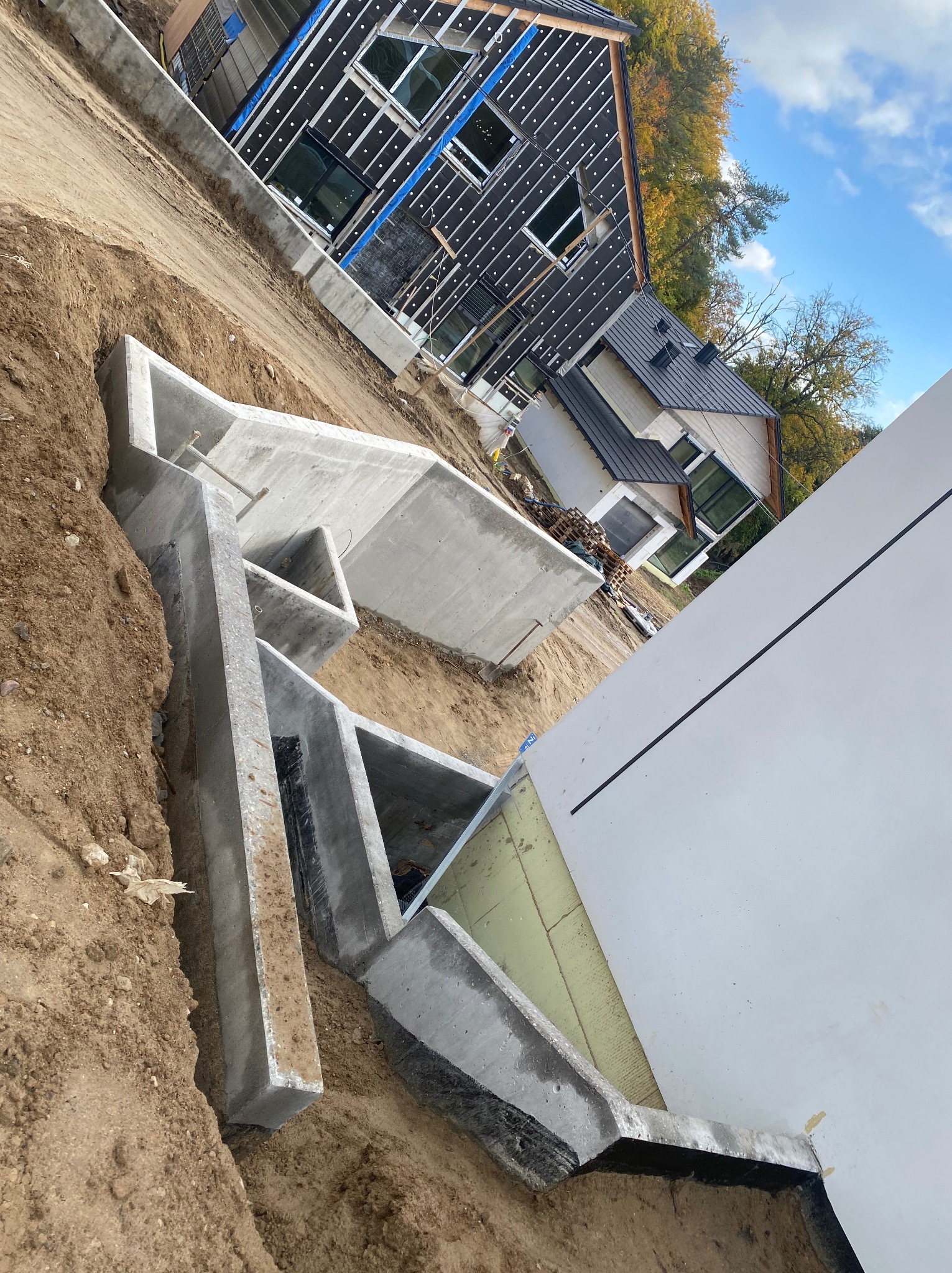 Budowa domu jednorodzinnego: fundamenty z betonu w wykopie, ściany z izolacją. Widoczne elementy konstrukcyjne i dom w tle, jesienne drzewa i błękitne niebo.