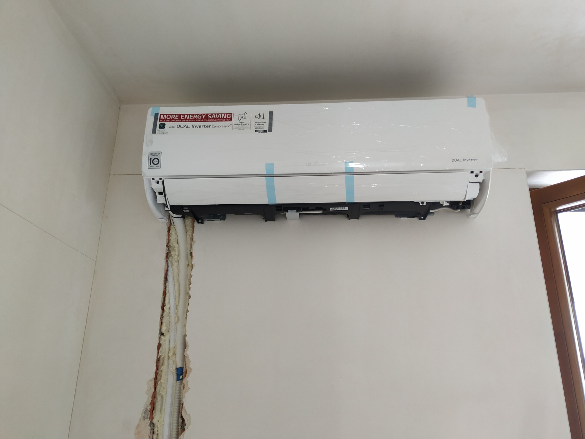 Nowo zamontowana jednostka wewnętrzna klimatyzacji LG, jeszcze w folii ochronnej i z nieusuniętymi taśmami, widoczne ślady pianki montażowej wokół przewodów w ścianie.