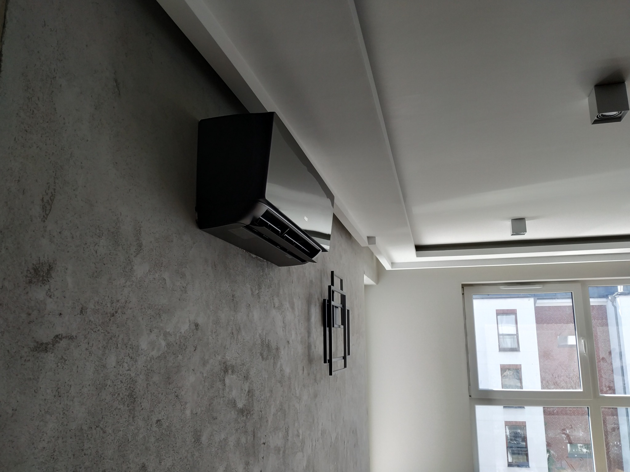 Czarny klimatyzator ścienny zamontowany w nowoczesnym wnętrzu z dekoracyjną ścianą o fakturze betonu i minimalistyczną ozdobą ścienną.