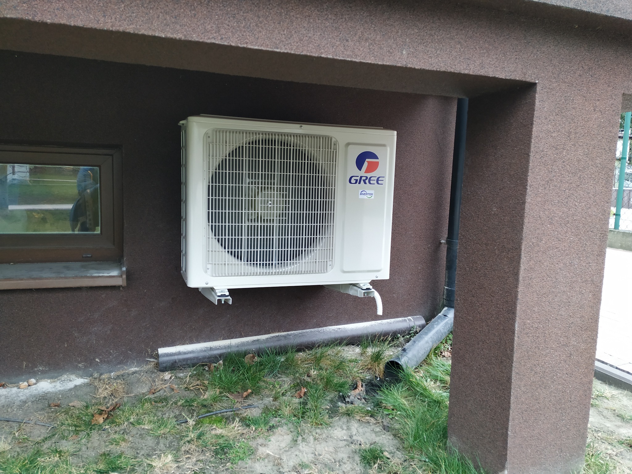 Zewnętrzna jednostka klimatyzacji GREE zamontowana na brązowej ścianie budynku, widoczna rura odprowadzająca skropliny i fragment trawnika.