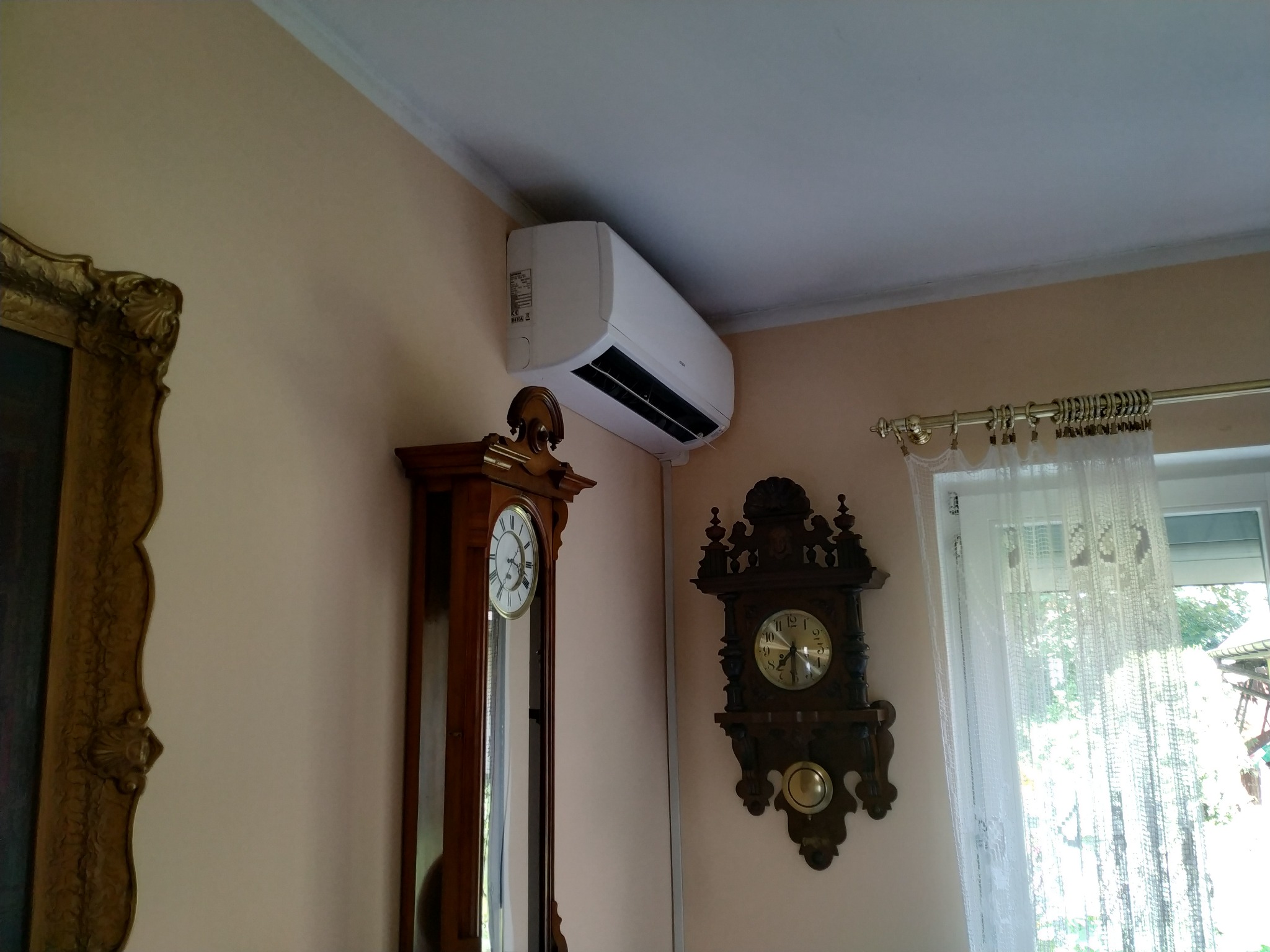 Biały klimatyzator ścienny zamontowany w pomieszczeniu z dwoma starymi zegarami i firanką w oknie.