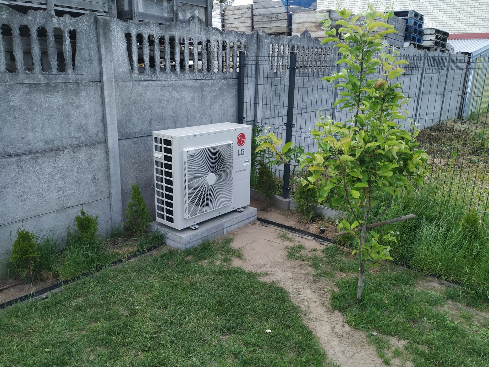 Zewnętrzna jednostka klimatyzacji LG umieszczona na betonowym fundamencie w ogrodzie, widok z boku, na tle betonowego i metalowego ogrodzenia.