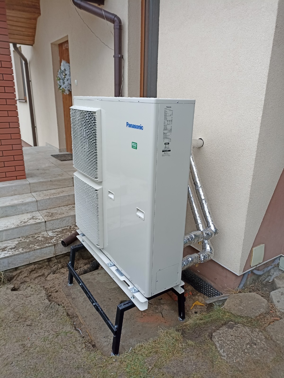 Biała pompa ciepła Panasonic R32 zamontowana na czarnej metalowej ramie na zewnątrz budynku z widocznymi rurami izolowanymi folią aluminiową.