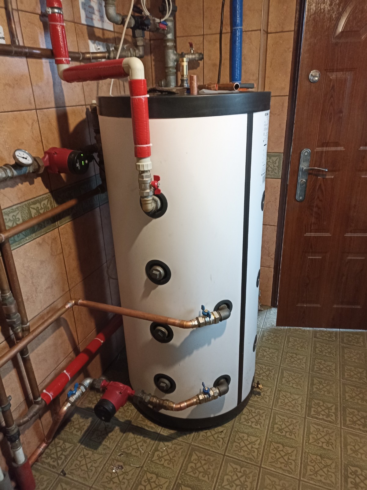 Biały zasobnik ciepłej wody użytkowej z licznymi przyłączami rur miedzianych i zaworów, widoczne pompy obiegowe, instalacja w pomieszczeniu z brązowymi płytkami.