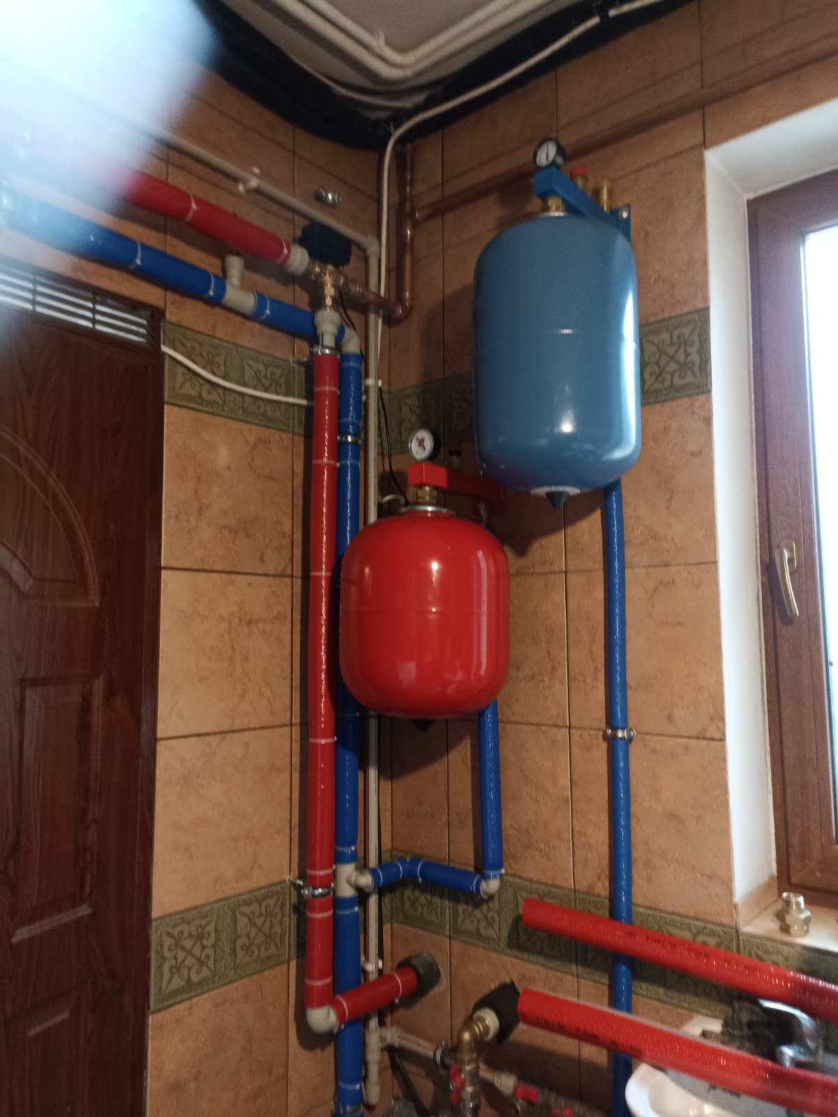 Czerwony i niebieski zbiornik wyrównawczy ciśnienia w instalacji CO, połączony rurami z manometrami, na tle brązowych płytek i drewnianych drzwi.