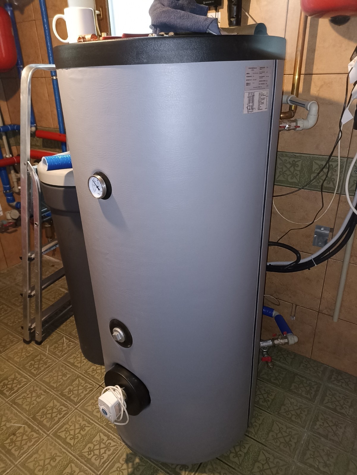 Szary, cylindryczny zbiornik z widocznym manometrem, elementami instalacji hydraulicznej i elektrycznej, na tle kafelkowanej ściany i podłogi.