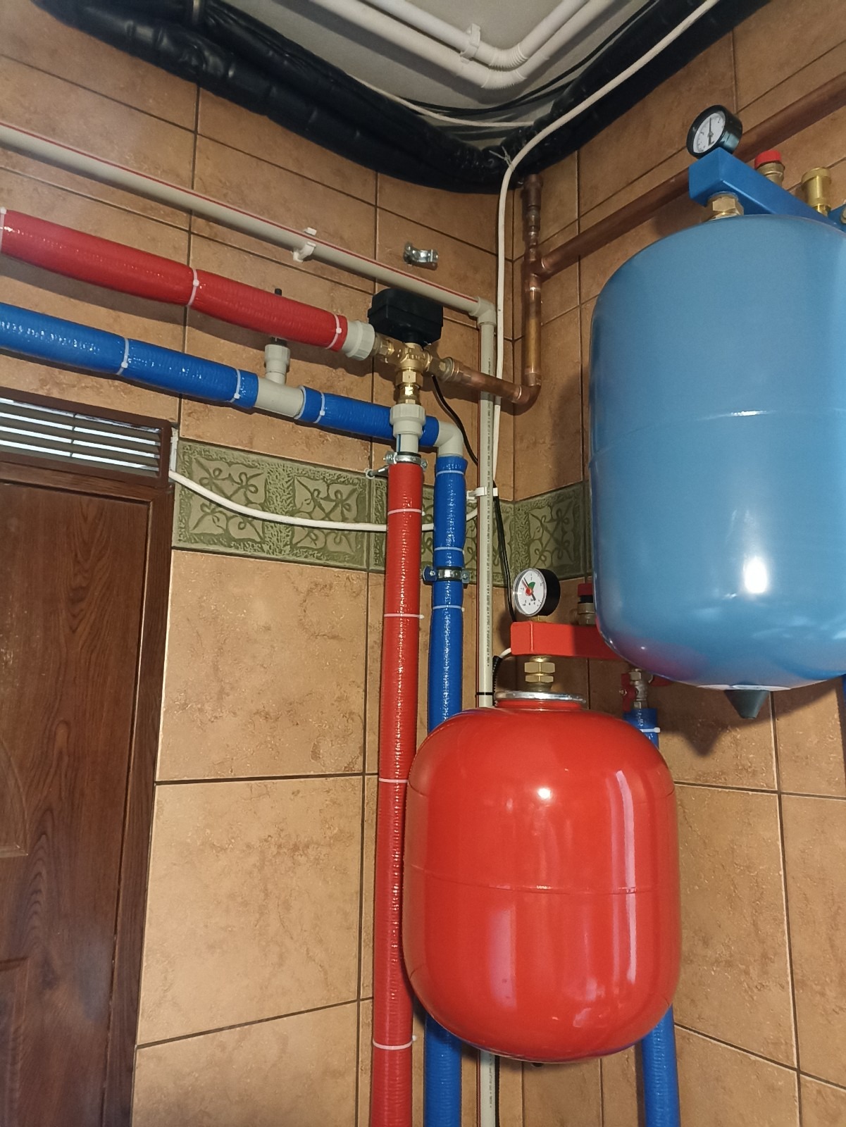 Instalacja hydrauliczna z czerwonym i niebieskim zbiornikiem wyrównawczym oraz rurami w izolacji, widoczny manometr i elementy miedziane, całość na tle brązowych płytek.