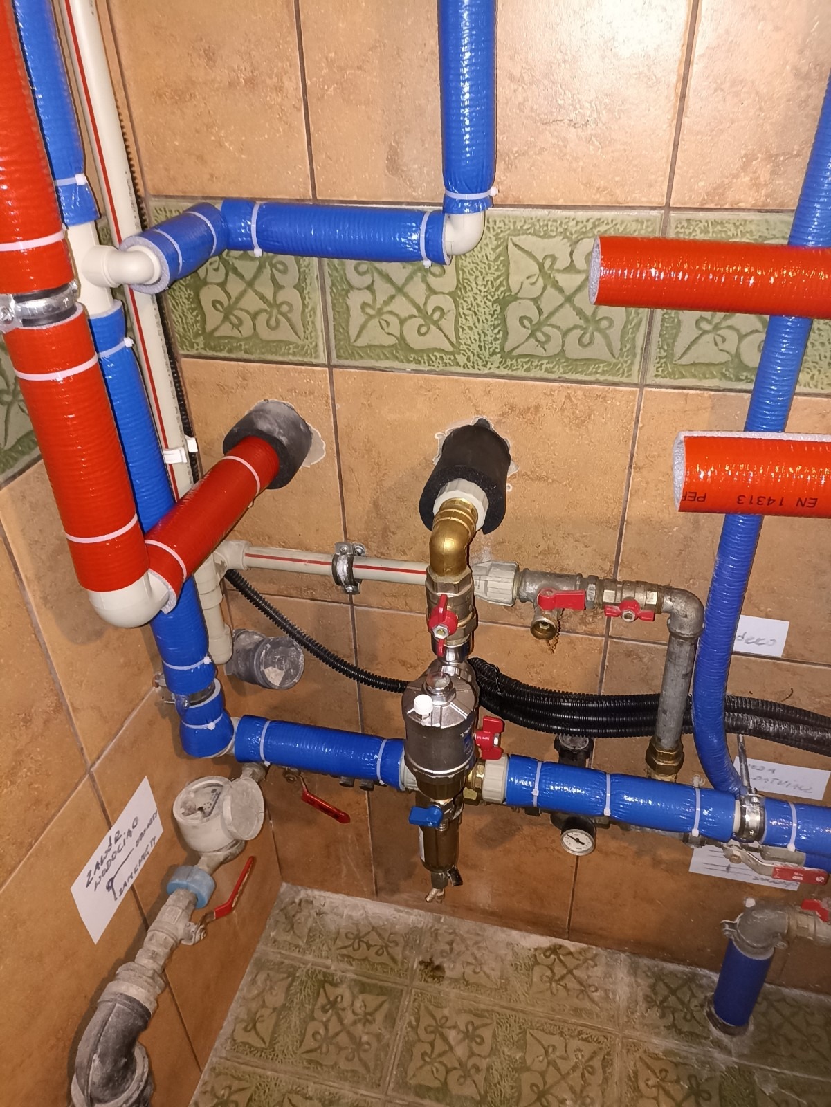 Widok z góry na skomplikowaną instalację hydrauliczną z rur czerwonych i niebieskich, zabezpieczonych folią, prowadzoną wzdłuż ściany z płytek ceramicznych i podłogi w łazience. Widoczny wodomierz...