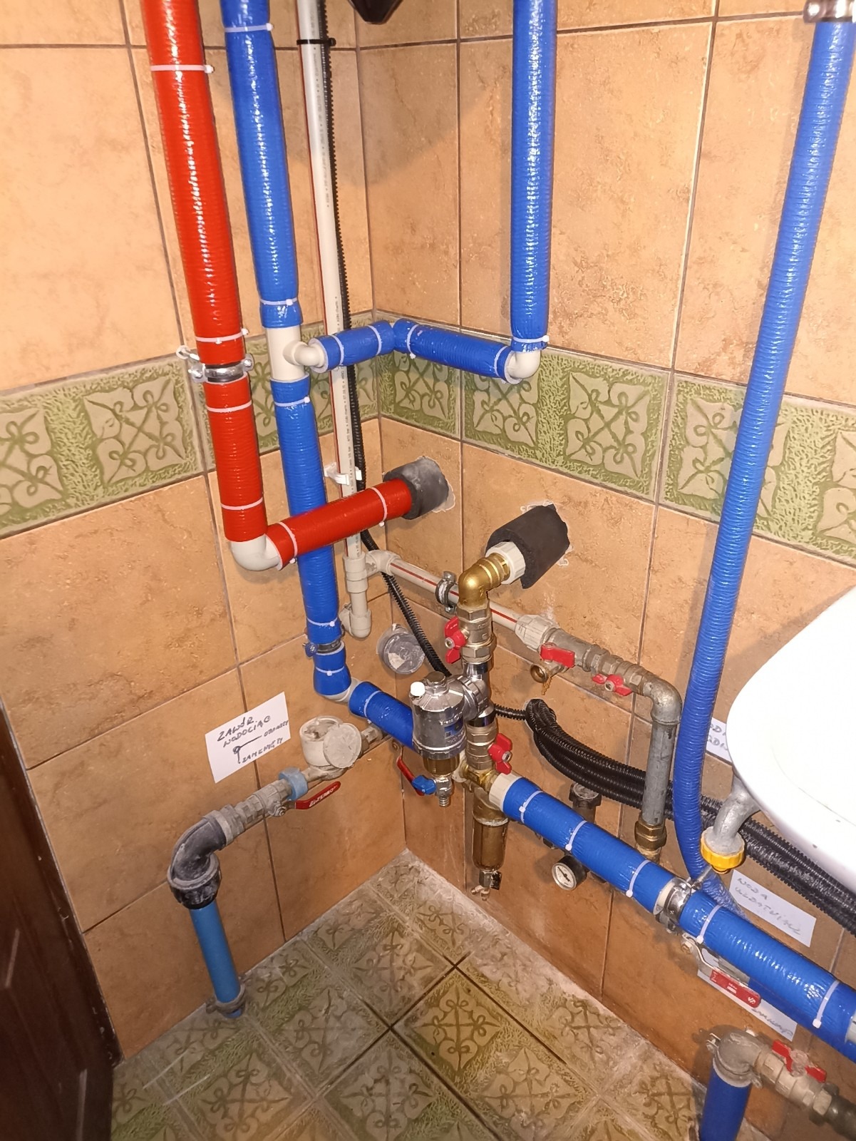 Złożona instalacja hydrauliczna z rurami w kolorach czerwonym i niebieskim, zabezpieczona białą taśmą, widoczne liczniki i zawory, zamontowana w pomieszczeniu z brązowymi płytkami.