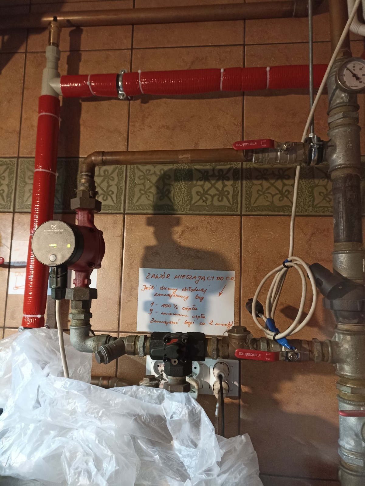 Fragment instalacji centralnego ogrzewania z pompą obiegową Grundfos, zaworami INSTALINE i izolowanymi rurami w kolorze czerwonym, uzupełniony o odręczną notatkę dotyczącą regulacji zaworu...