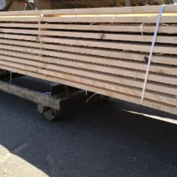 Stos świeżo przetartej tarcicy sosnowej, ułożonej na wózku transportowym z metalową ramą i kołami, zabezpieczonej białą taśmą, widoczny cień na betonowym podłożu.