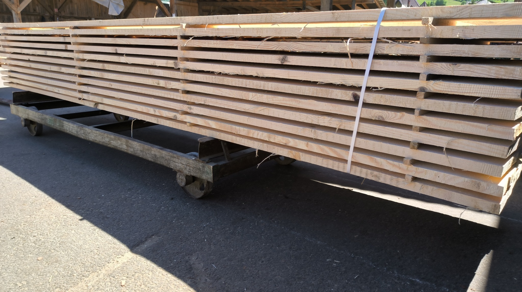 Stos świeżo przetartej tarcicy sosnowej, ułożonej na wózku transportowym z metalową ramą i kołami, zabezpieczonej białą taśmą, widoczny cień na betonowym podłożu.