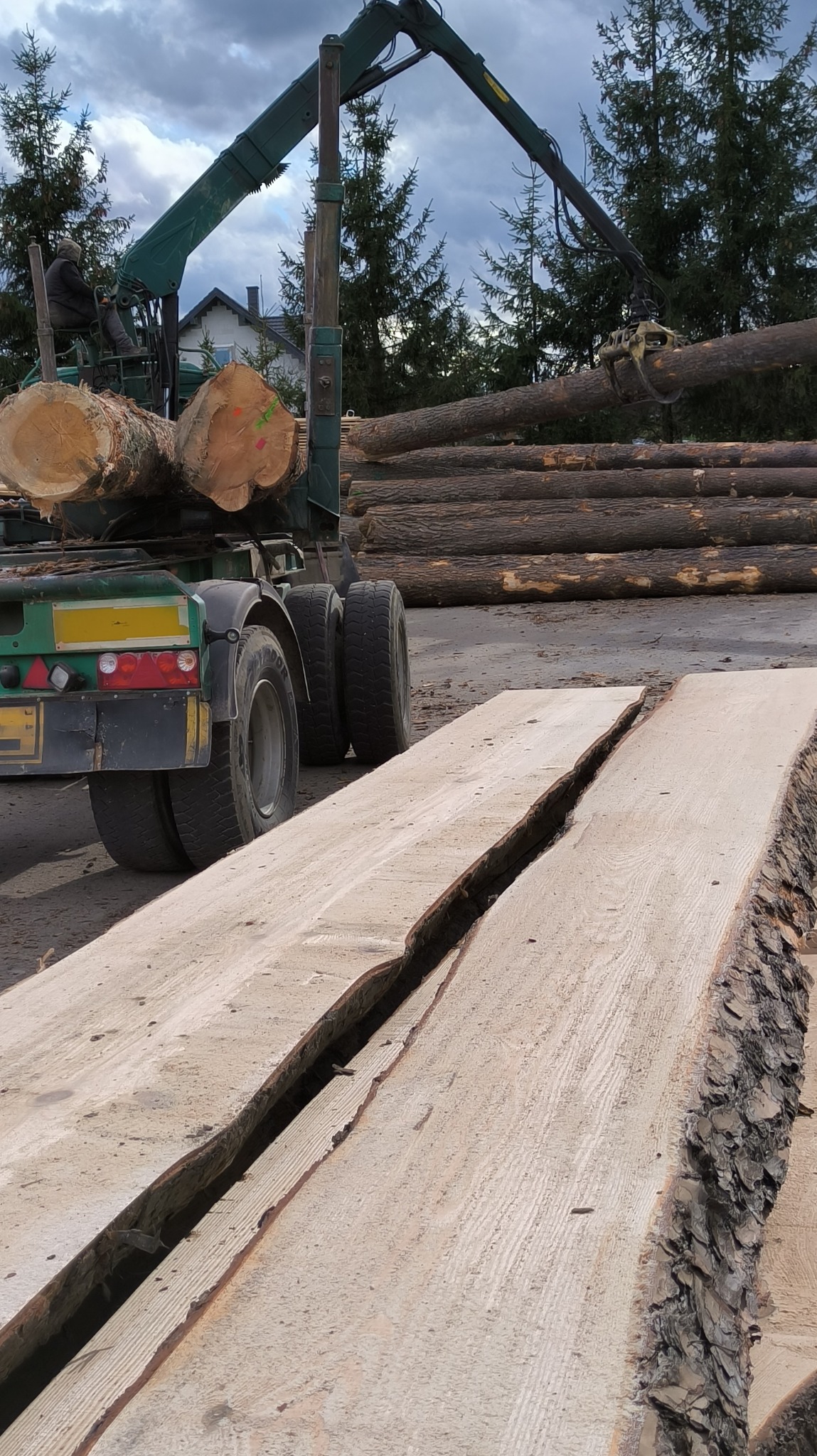 Załadunek kłód drewna na przyczepę z wykorzystaniem dźwigu, na pierwszym planie dwie grube deski z korą na krawędziach.
