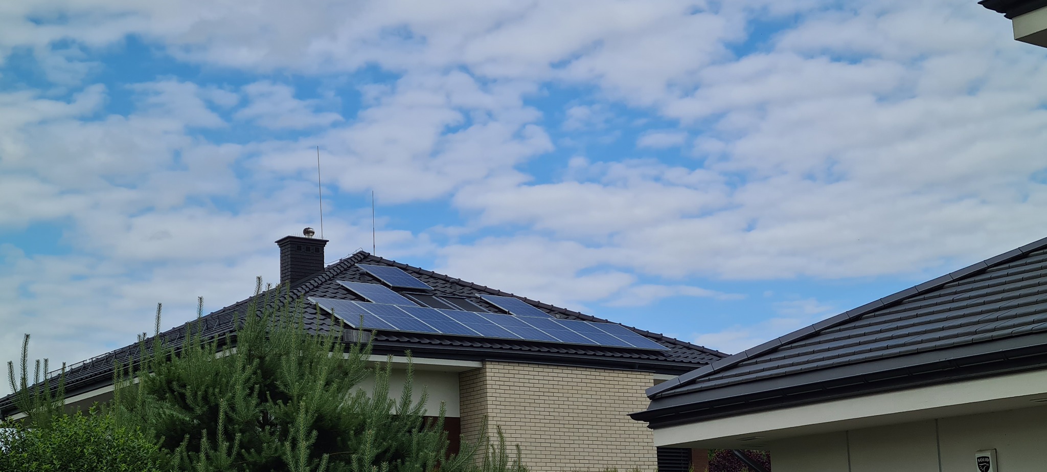 Panele słoneczne na dachu domu jednorodzinnego w Dmosinie, widok z poziomu ogrodu na tle błękitnego nieba z obłokami.