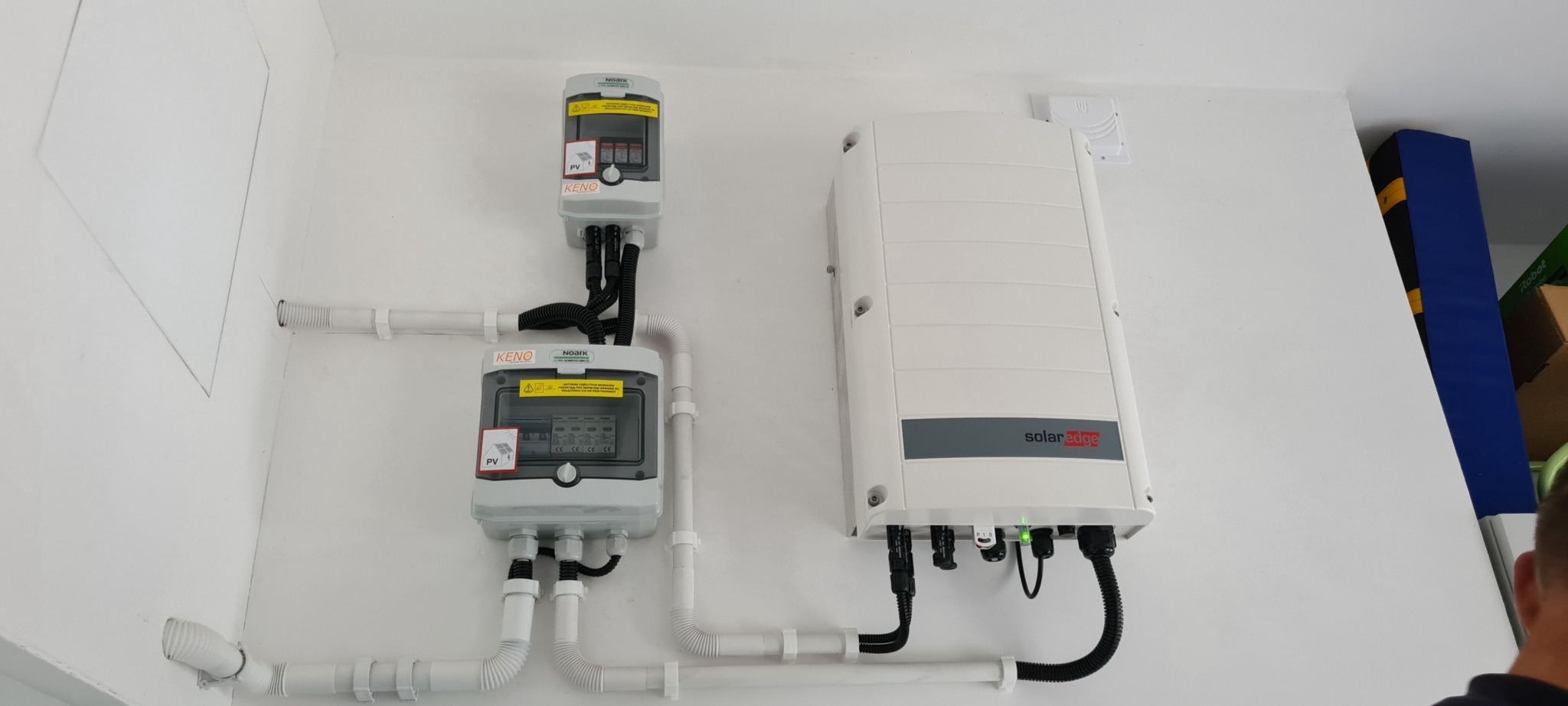 Dwa rozdzielacze elektryczne Noark Keno i falownik SolarEdge zamontowane na białej ścianie, połączone białymi rurami PCV i czarnymi kablami.