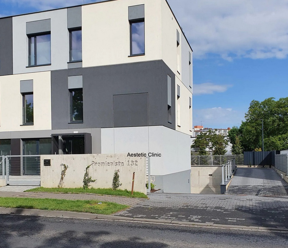 Fasada nowoczesnego budynku Aesthetic Clinic z betonowym ogrodzeniem, na którym widnieje adres Promienista 132, z zielonymi pnączami u podstawy.