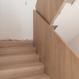 WoodCare Sp. z o.o. - Jasne, drewniane schody z minimalistyczną balustradą, widoczne ślady prac wykończeniowych na ścianie. Ujęcie z dołu, podkreślające naturalny odcień drewna.