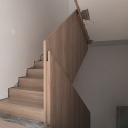 WoodCare Sp. z o.o. - Drewniane schody z balustradą w trakcie montażu, widoczne niedokończone wykończenia ścian i podłogi. Ujęcie z dołu, z widocznym logo firmy na dole kadru.