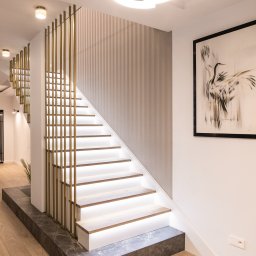 WoodCare Sp. z o.o. - Nowoczesne schody z podświetleniem LED i złotą balustradą w eleganckim wnętrzu. Widoczny fragment parkietu i obraz na ścianie. Minimalistyczny design.