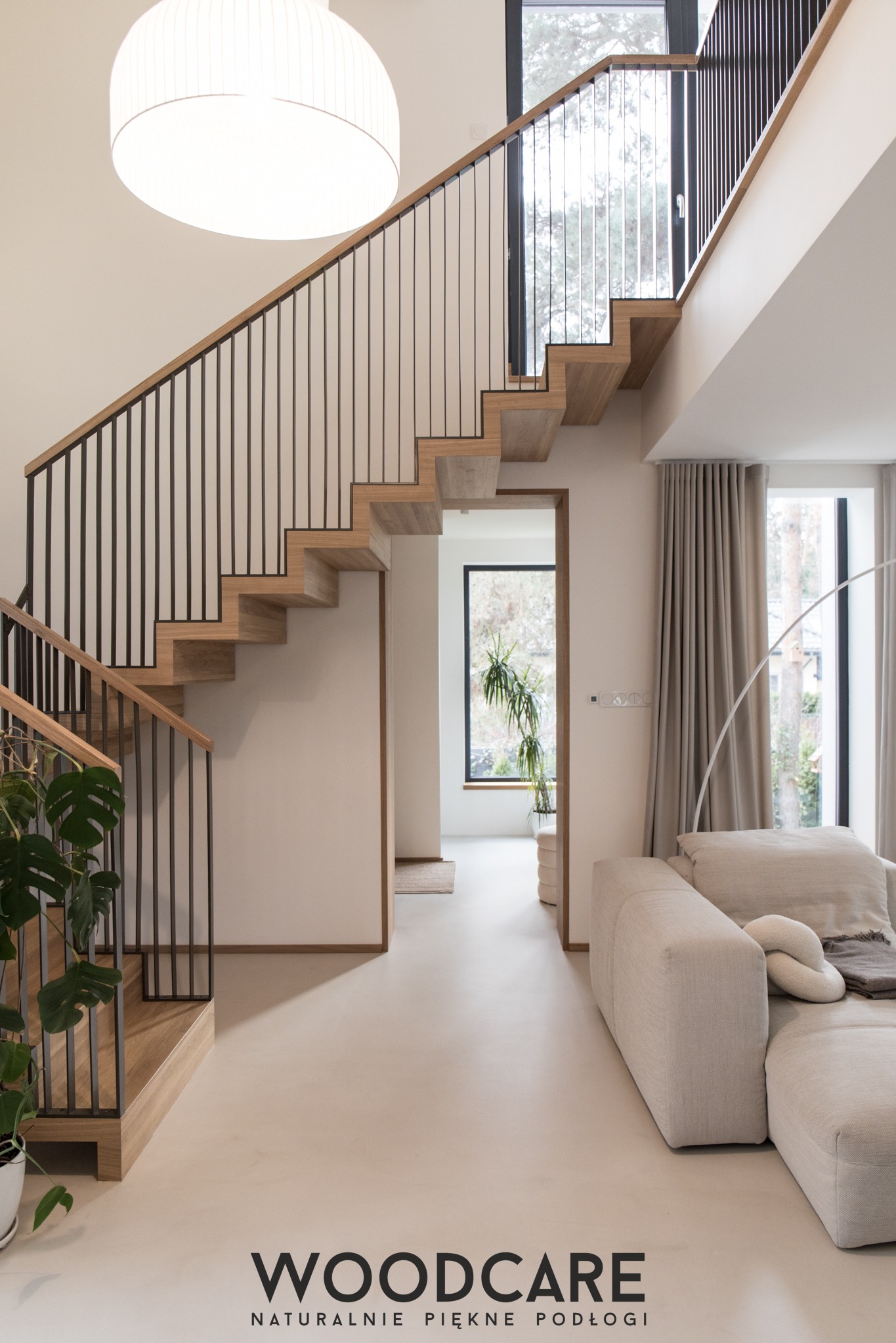 Nowoczesne schody z drewnianymi stopniami i czarną balustradą w jasnym wnętrzu. Minimalistyczny design, duże okna, naturalne światło. Widok na salon z sofą.
