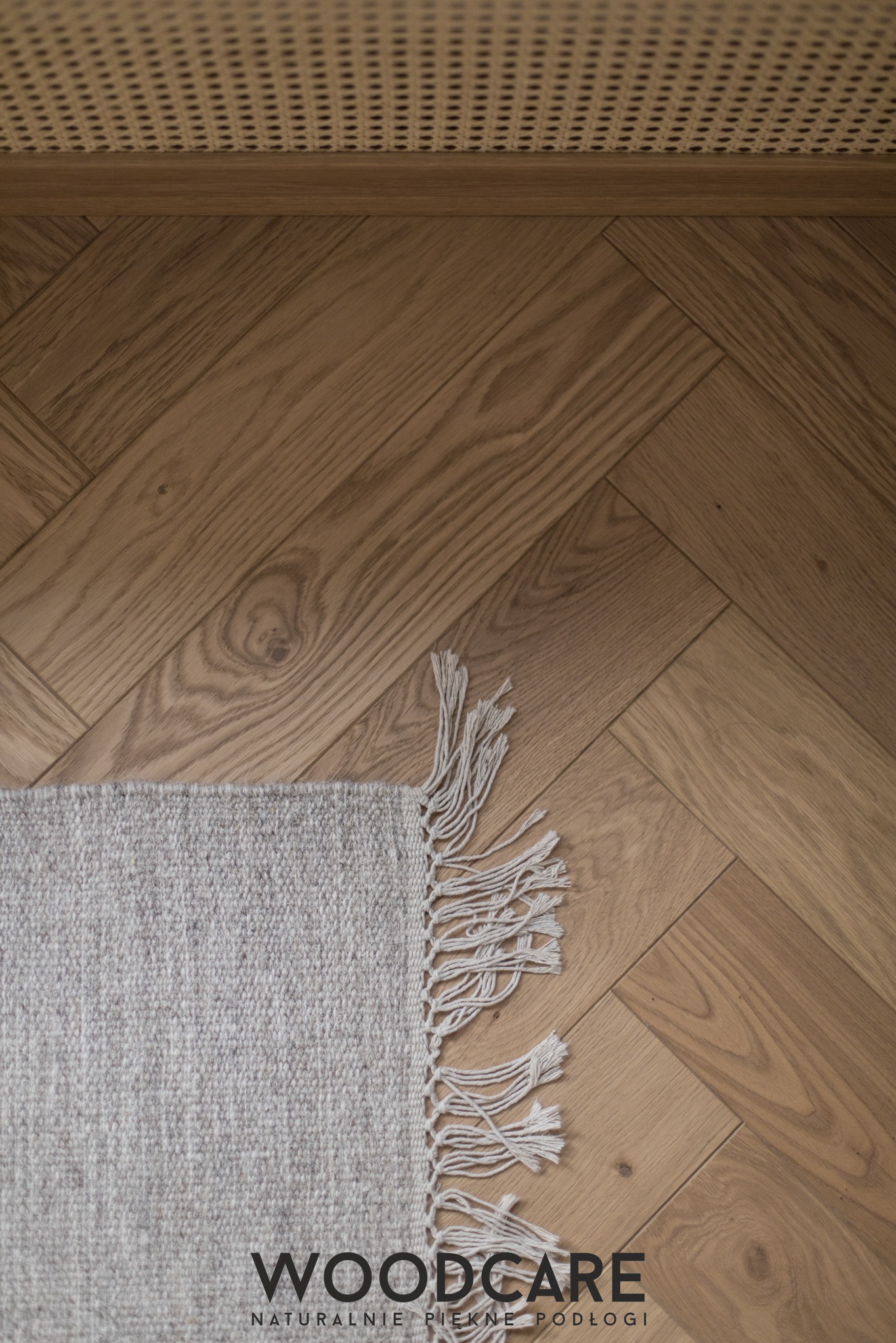 Parkiet ułożony w jodełkę francuską, z widocznym fragmentem dywanu z frędzlami i kratką wentylacyjną. Ciepłe odcienie drewna i naturalne światło.