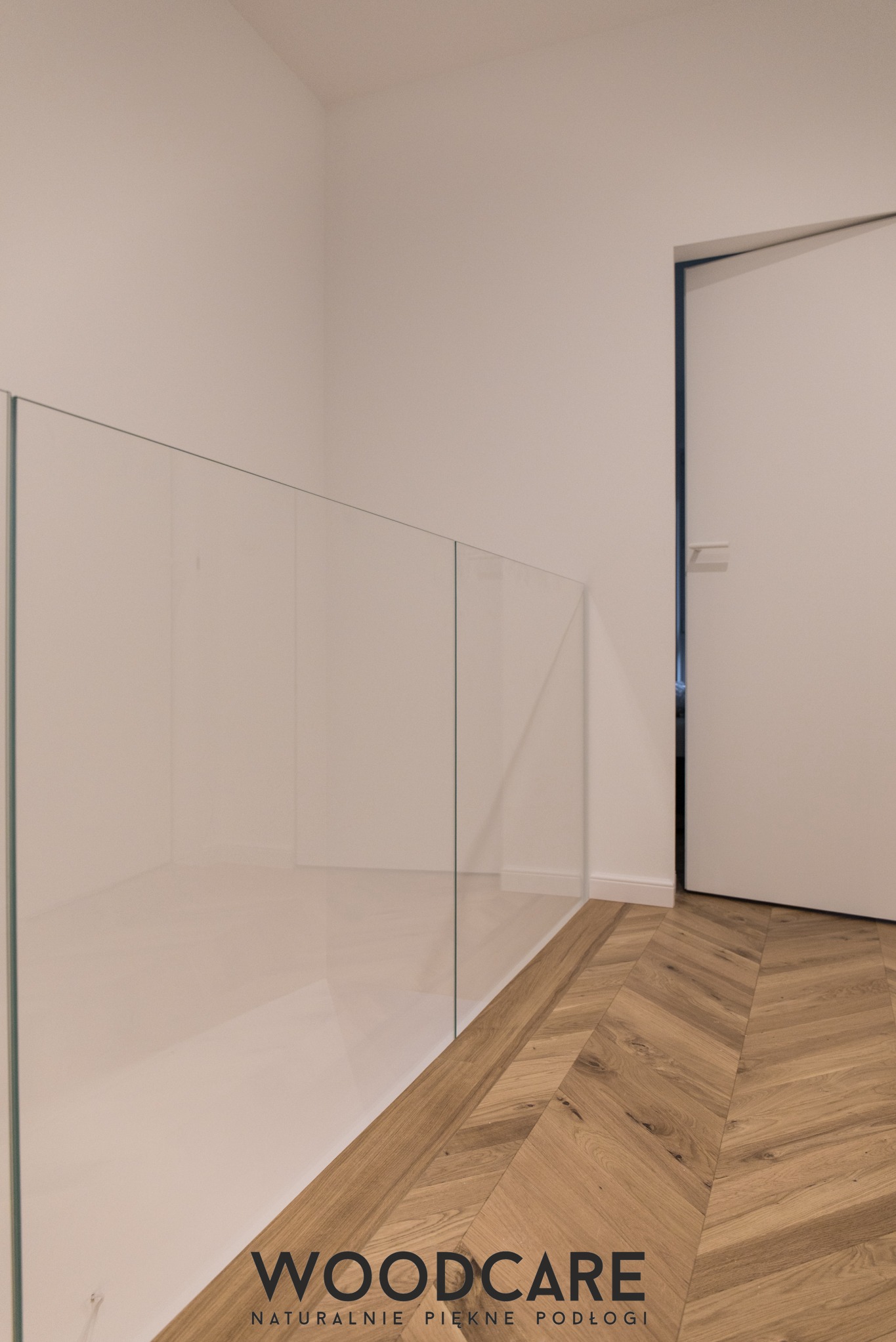 Jasne wnętrze z parkietem ułożonym we wzór jodełki, szklana balustrada i białe drzwi. Widoczny fragment logo firmy Woodcare.
