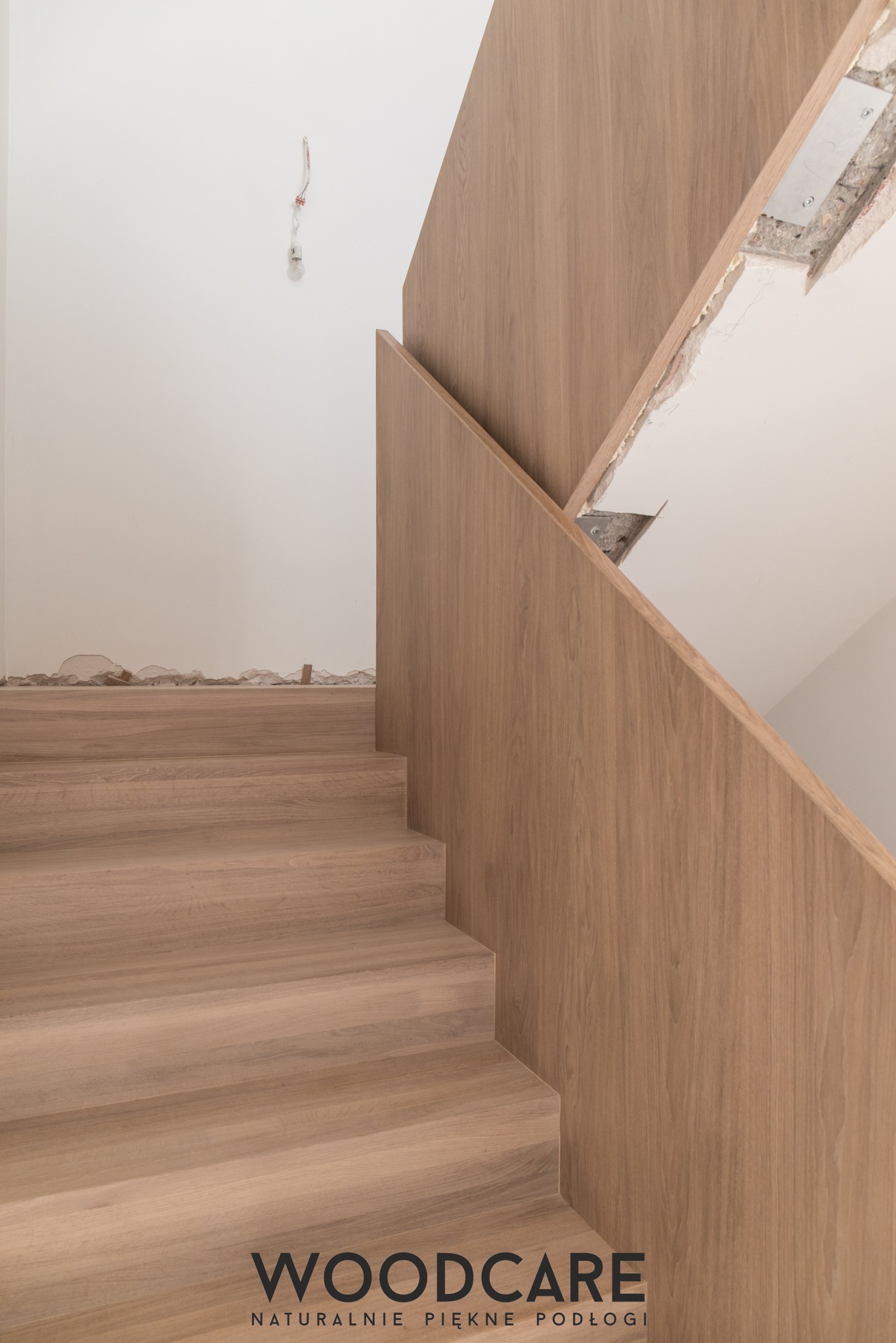 Jasne, drewniane schody z minimalistyczną balustradą, widoczne ślady prac wykończeniowych na ścianie. Ujęcie z dołu, podkreślające naturalny odcień drewna.