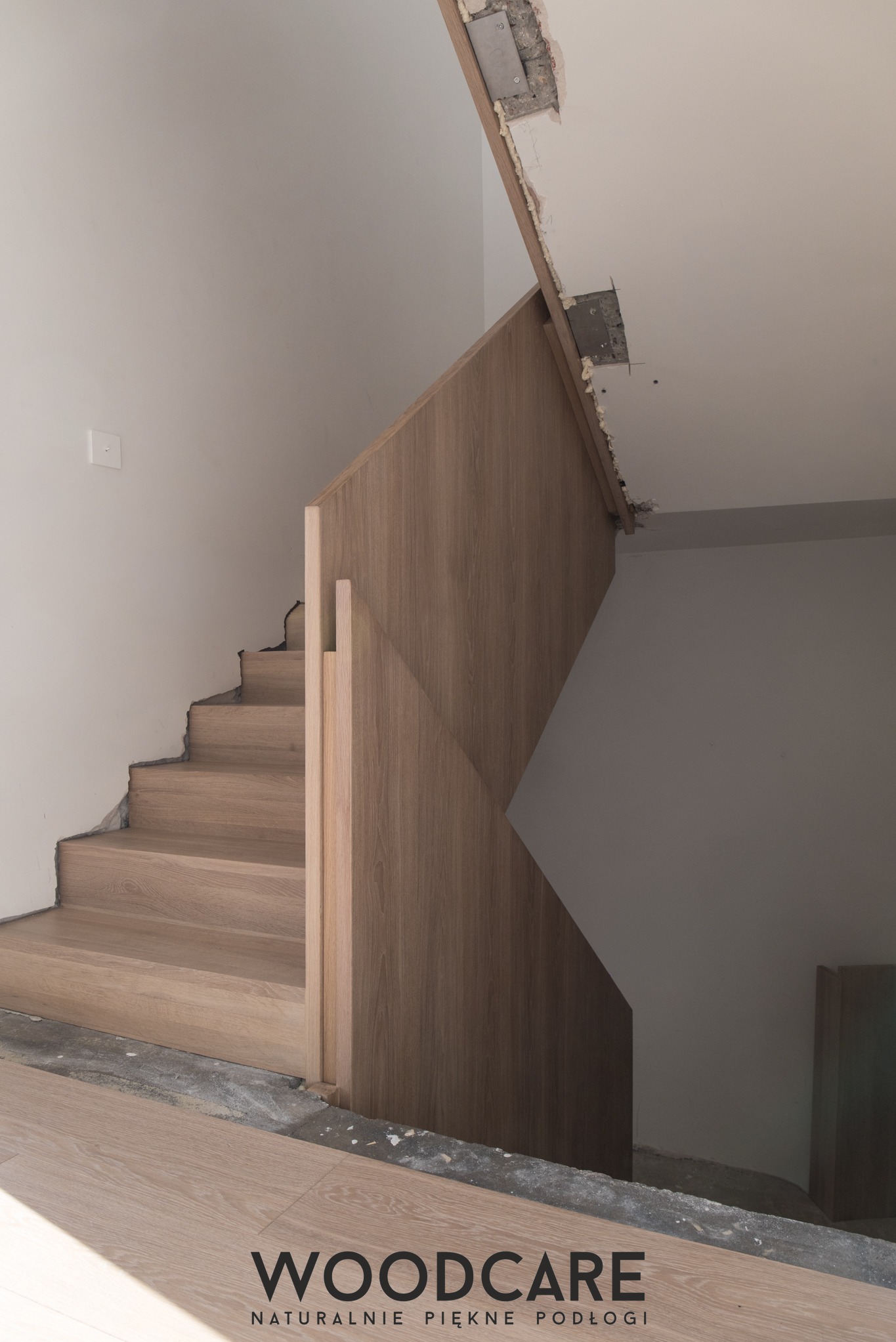Drewniane schody z balustradą w trakcie montażu, widoczne niedokończone wykończenia ścian i podłogi. Ujęcie z dołu, z widocznym logo firmy na dole kadru.