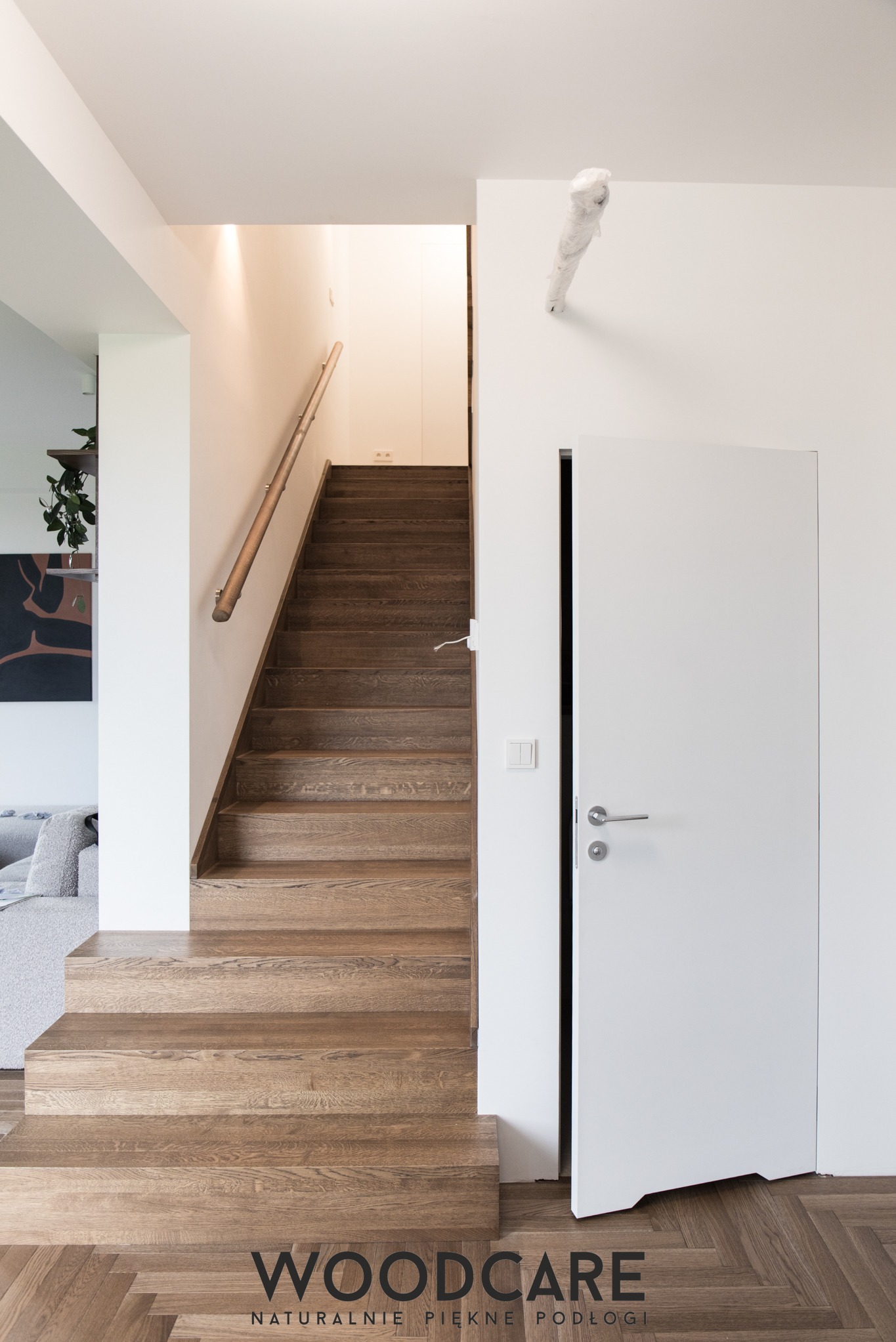 Jasne, drewniane schody z poręczą i białe drzwi wewnętrzne w nowoczesnym wnętrzu domu w Warszawie. Parkiet ułożony w jodełkę. Minimalistyczny design.