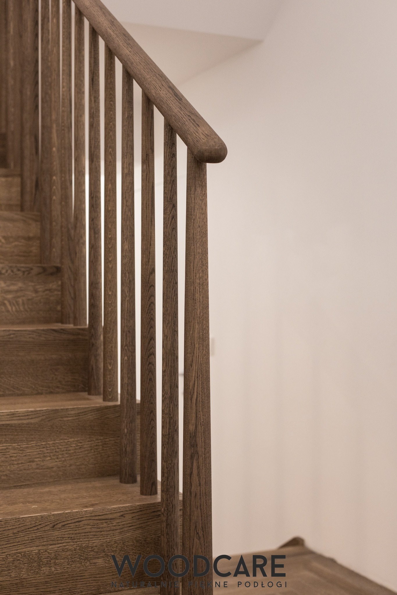 Detal drewnianych schodów z balustradą w odcieniach brązu, widok z dołu, z bliska. Minimalistyczny design, naturalne wykończenie drewna, jasne tło ściany.