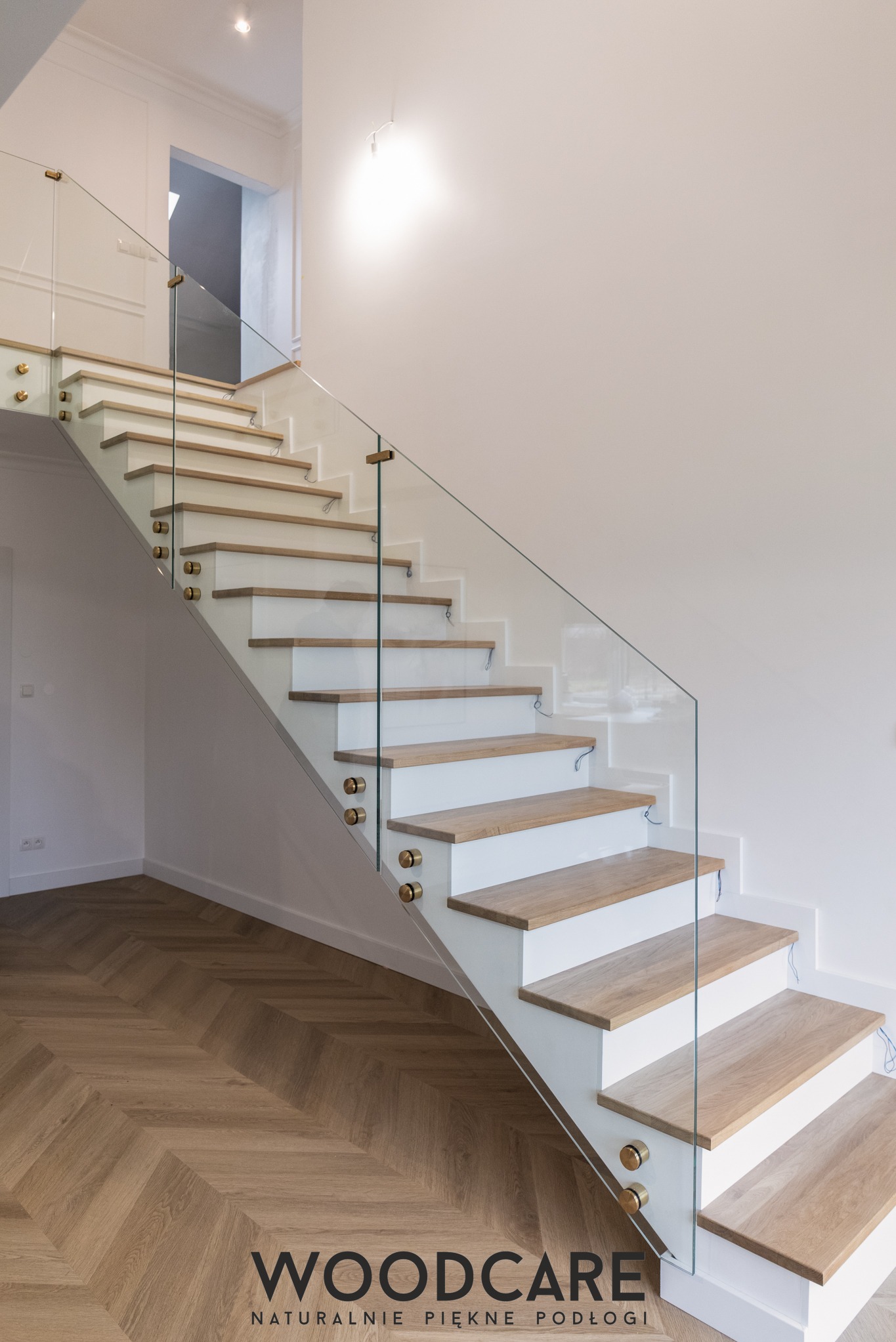 Nowoczesne schody z drewnianymi stopniami i szklaną balustradą, złote mocowania. Parkiet ułożony w jodełkę. Minimalistyczne wnętrze z białymi ścianami i subtelnym oświetleniem.